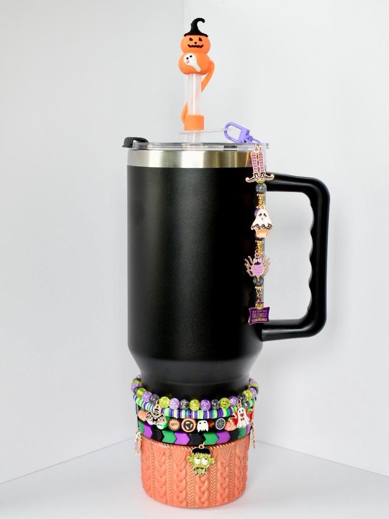 Black tumbler with a handle, adorned with Halloween-themed charms and a decorative sleeve. The sleeve is orange with a cable knit design. A series of beaded bracelets in purple, green, black, and orange encircle the base. A pumpkin-shaped straw topper sits on top, adding a playful touch.