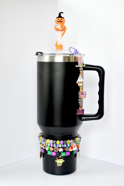 Black tumbler with a silver rim, black handle, and lid. A Halloween-themed charm hangs from the handle. The tumbler is decorated with multiple bracelets in green, purple, and black. A Halloween-themed straw topper sits on top.