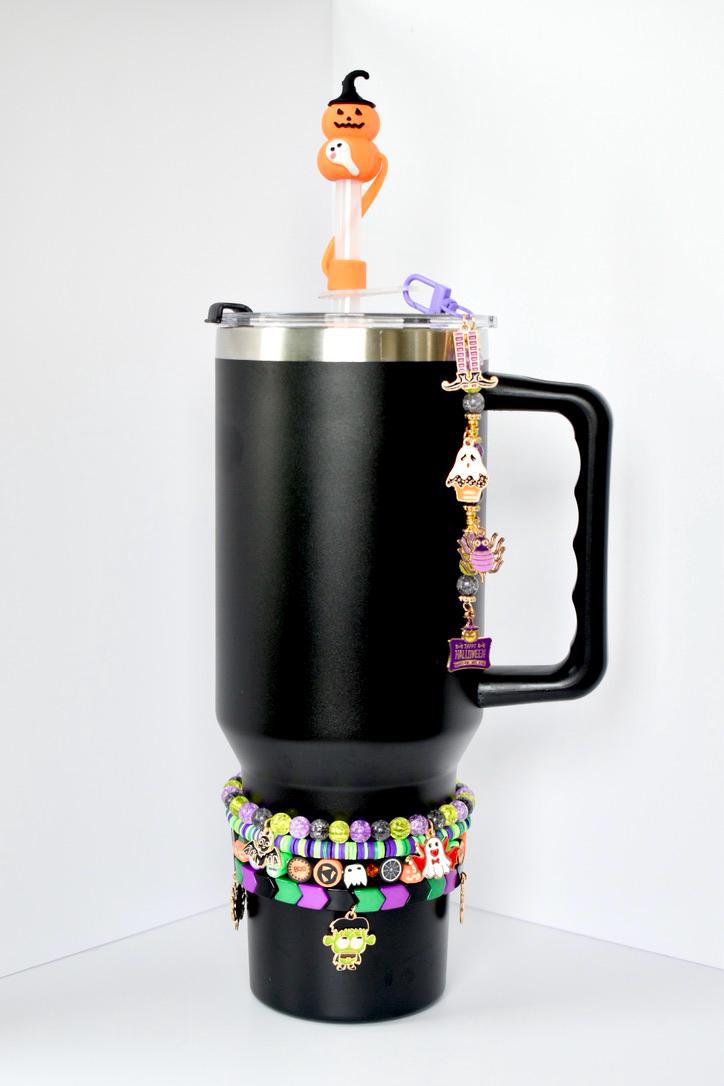 Black tumbler with a silver rim, black handle, and lid. A Halloween-themed charm hangs from the handle. The tumbler is decorated with multiple bracelets in green, purple, and black. A Halloween-themed straw topper sits on top.