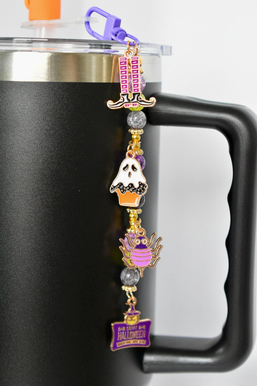 A Halloween-themed charm hanging from a purple clasp attached to a black mug. The charm features a witch's hat, a ghost in an orange cupcake, a purple spider, and a sign that reads 'Happy Halloween'. The charms are gold and purple with gray and gold beads.
