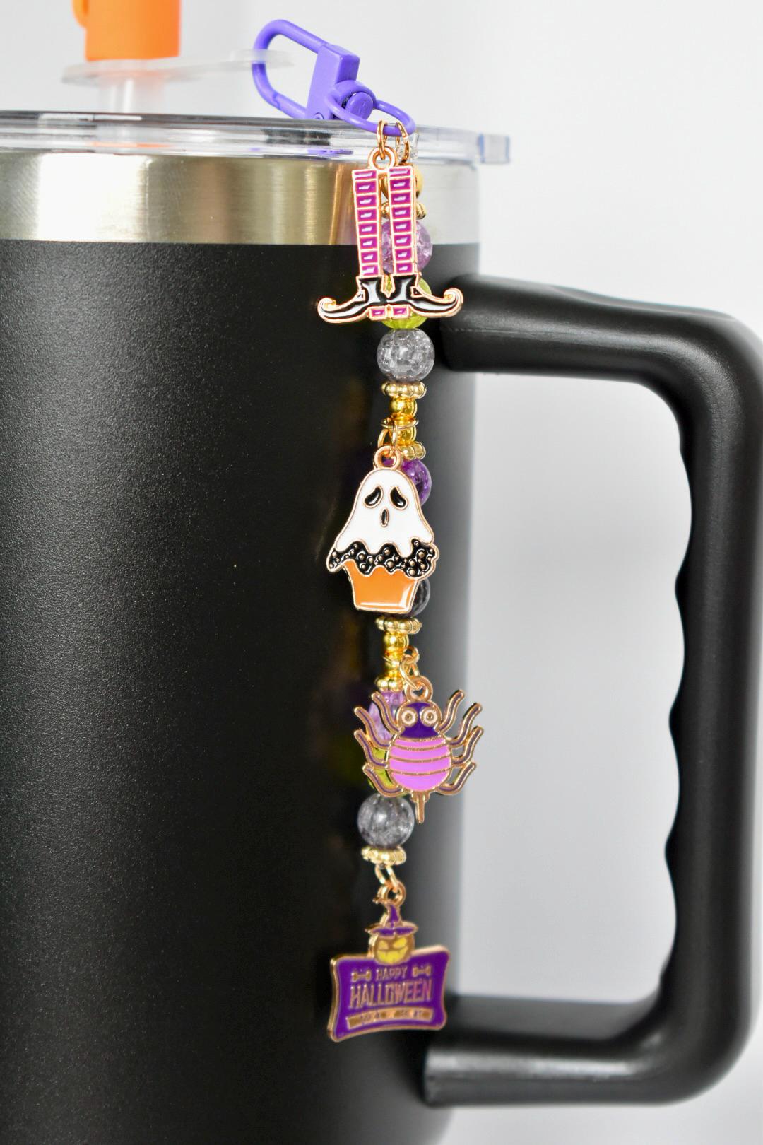 A Halloween-themed charm hanging from a purple clasp attached to a black mug. The charm features a witch's hat, a ghost in an orange cupcake, a purple spider, and a sign that reads 'Happy Halloween'. The charms are gold and purple with gray and gold beads.