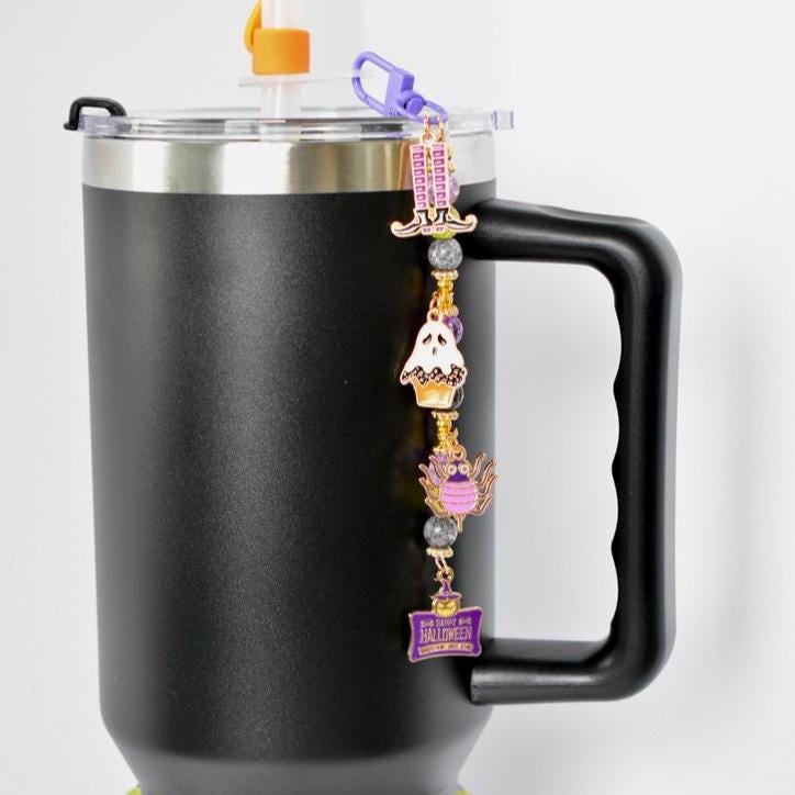 A black insulated travel mug with a silver rim and handle. A purple charm is attached to the lid, featuring Halloween-themed charms: a tombstone, a ghost, a cupcake, and a purple owl. The charm has a purple tag that reads 'Happy Halloween'.