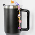 A black insulated travel mug with a silver rim and handle. A purple charm is attached to the lid, featuring Halloween-themed charms: a tombstone, a ghost, a cupcake, and a purple owl. The charm has a purple tag that reads 'Happy Halloween'.