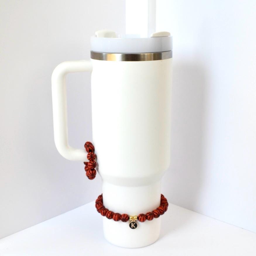 A white insulated tumbler with a handle, a silver rim, and a white lid. Two red and brown basketball-themed bracelets are attached to the tumbler. One bracelet is around the handle, and the other is around the base of the tumbler.