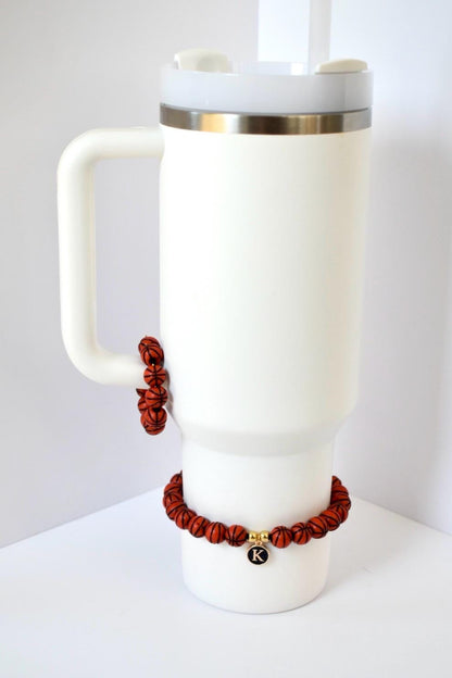 A white insulated tumbler with a handle and a silver rim, adorned with two bracelets made of small, orange basketball-shaped beads. One bracelet is attached to the handle, and the other is around the base. A gold charm with the letter "K" hangs from the lower bracelet.