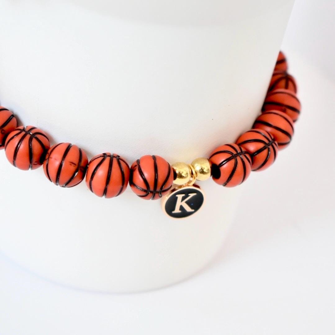 A beaded bracelet featuring orange basketball-shaped beads with black lines. The bracelet includes gold-colored beads and a charm with the letter "K" on a black background. The bracelet is displayed on a white surface.