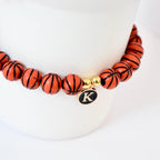 A beaded bracelet featuring orange basketball-shaped beads with black lines. The bracelet includes gold-colored beads and a charm with the letter "K" on a black background. The bracelet is displayed on a white surface.