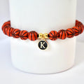 A basketball-themed bracelet featuring orange beads with black lines, resembling basketballs. The bracelet includes a gold-colored charm with the letter "K" on a black background. The bracelet is displayed on a white background.
