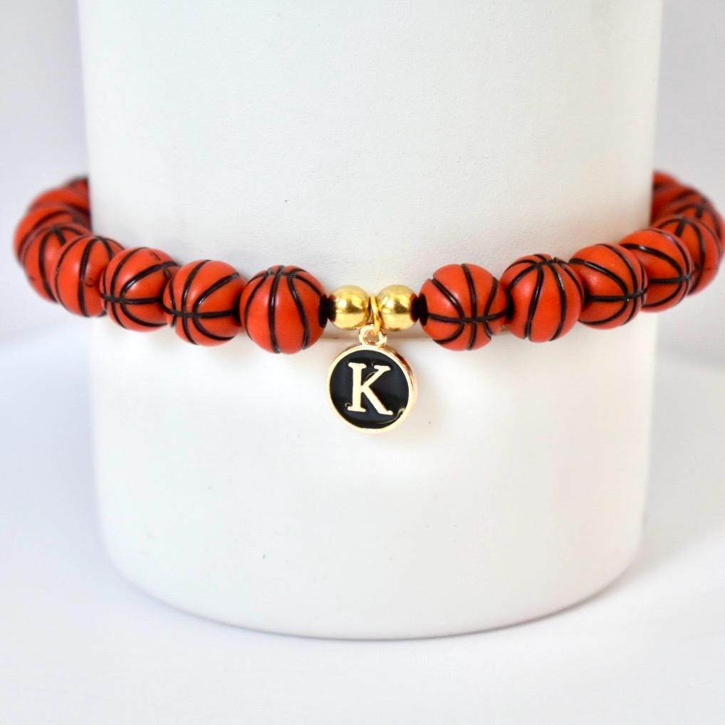A basketball-themed bracelet featuring orange beads with black lines, resembling basketballs. The bracelet includes a gold-colored charm with the letter "K" on a black background. The bracelet is displayed on a white background.