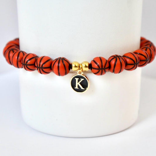 A basketball-themed bracelet featuring orange beads with black lines, resembling basketballs. The bracelet includes a gold-colored charm with the letter "K" on a black background. The bracelet is displayed on a white background.