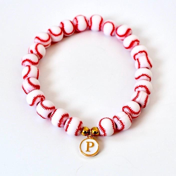 A baseball-themed bracelet featuring white beads with red stitching, resembling miniature baseballs. The bracelet has a gold-colored charm with the letter "P" in white. The bracelet is on a white background.
