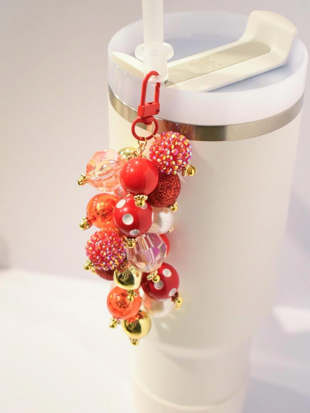 A charm for a tumbler cup featuring a cluster of colorful beads. The charm has a red lobster clasp and a gold chain. The beads are various sizes and colors, including red, gold, and clear. Some beads are glittery or have white polka dots. The charm is attached to a white tumbler with a straw.