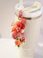 A charm for a tumbler cup featuring a cluster of colorful beads. The charm has a red lobster clasp and a gold chain. The beads are various sizes and colors, including red, gold, and clear. Some beads are glittery or have white polka dots. The charm is attached to a white tumbler with a straw.