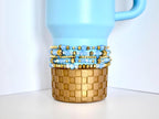 Light blue and gold tumbler bracelet set with iridescent beads and gold spacers, displayed on a soft blue tumbler with a checker-patterned gold base.