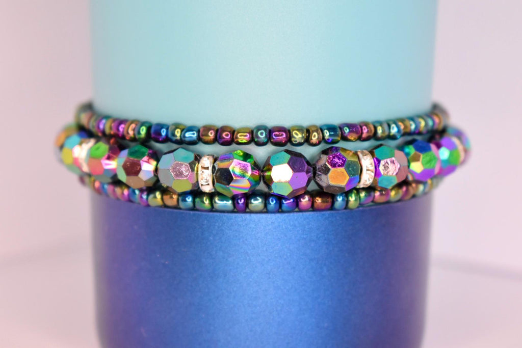 Colorful iridescent beaded tumbler bracelets with shiny metallic hues and rhinestone accents, wrapped around a two-tone blue tumbler.