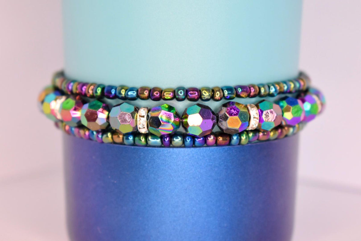 Colorful iridescent beaded tumbler bracelets with shiny metallic hues and rhinestone accents, wrapped around a two-tone blue tumbler.