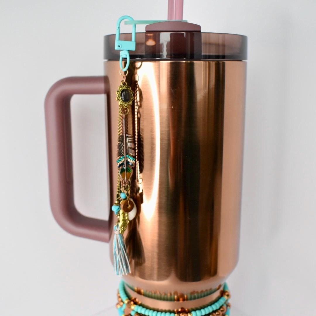 A copper-colored insulated tumbler with a handle, a teal clip, and a decorative charm. The charm features feathers, beads, and a tassel. A turquoise beaded bracelet is wrapped around the base of the tumbler. A pink straw is inserted into the lid.