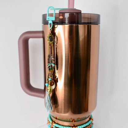 A copper-colored insulated tumbler with a handle, a teal clip, and a decorative charm. The charm features feathers, beads, and a tassel. A turquoise beaded bracelet is wrapped around the base of the tumbler. A pink straw is inserted into the lid.