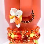 Pumpkin-patterned bow charm with a golden yellow center hanging from a bracelet made of shiny bronze beads, wrapped around the handle of a coral-colored Stanley tumbler.