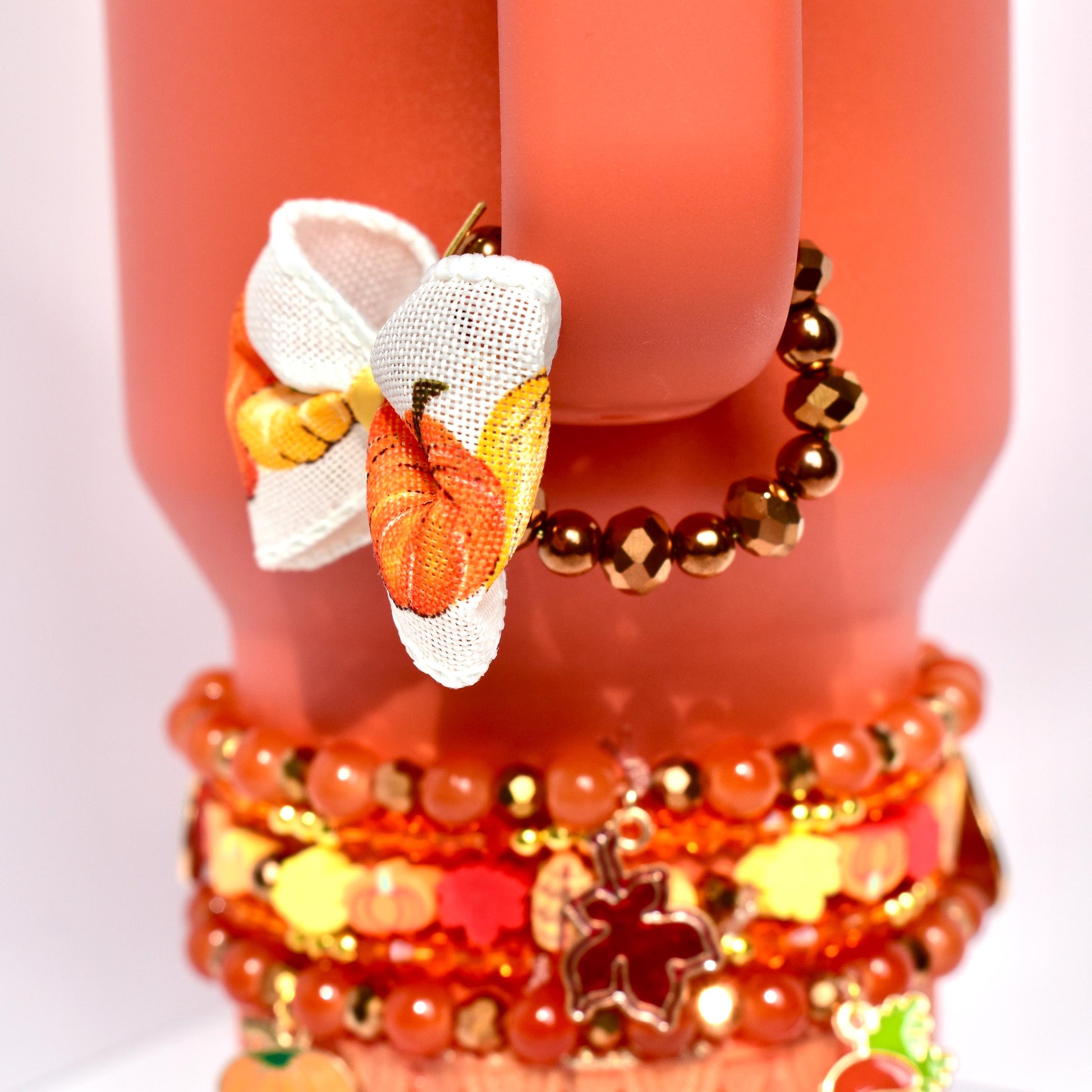 Pumpkin-patterned bow charm with a golden yellow center hanging from a bracelet made of shiny bronze beads, wrapped around the handle of a coral-colored Stanley tumbler.
