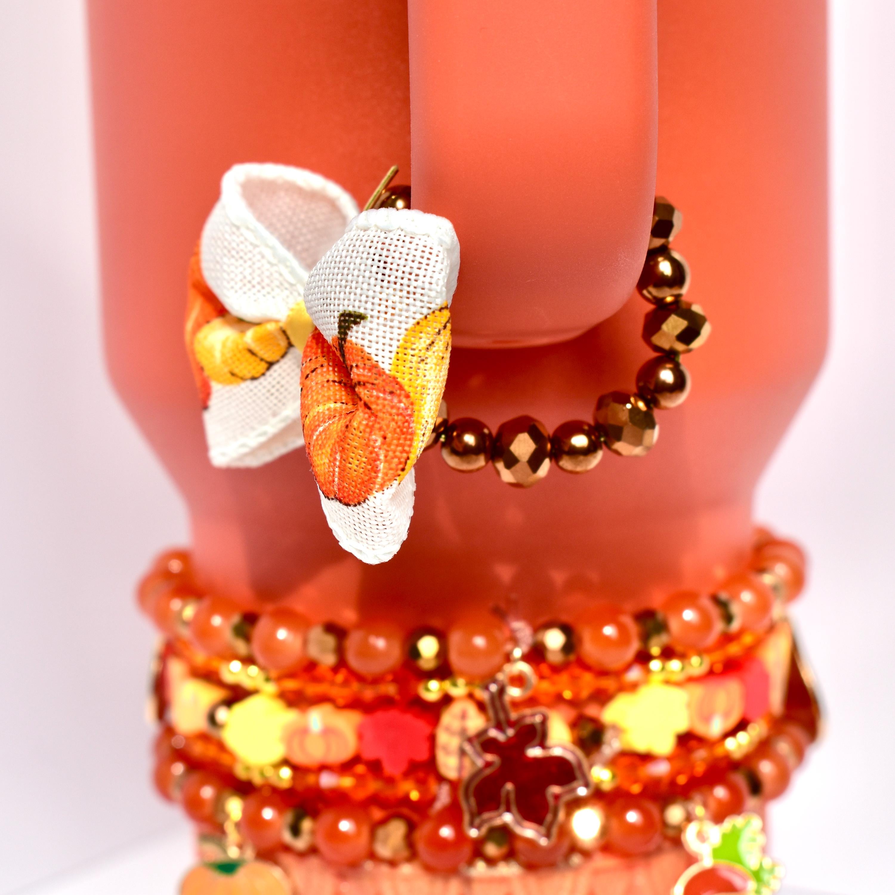 Pumpkin-patterned bow charm with a golden yellow center hanging from a bracelet made of shiny bronze beads, wrapped around the handle of a coral-colored Stanley tumbler.