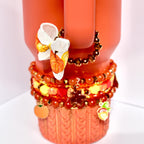 Autumn-inspired handle bracelet adorned with warm-toned beads and a small bow printed with vibrant orange and yellow pumpkins, clipped to the handle of a seasonal orange tumbler.