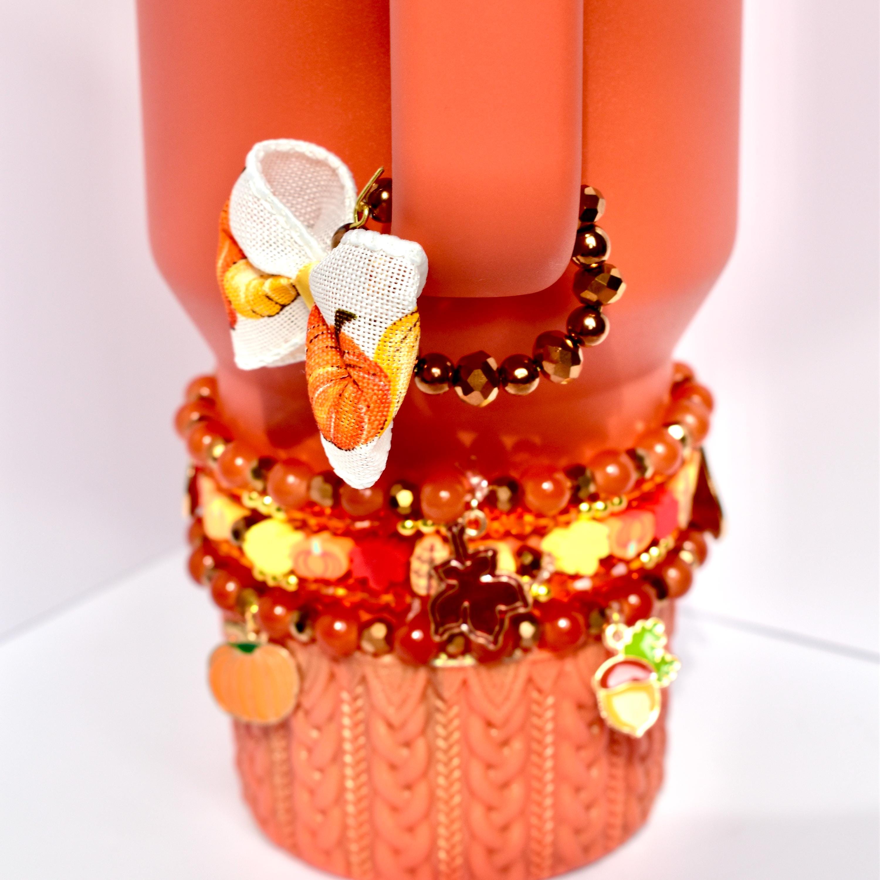 Autumn-inspired handle bracelet adorned with warm-toned beads and a small bow printed with vibrant orange and yellow pumpkins, clipped to the handle of a seasonal orange tumbler.