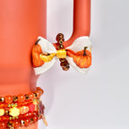 Fall tumbler handle bracelet made of glossy bronze and gold beads, accented with a mini bow showcasing an autumn pumpkin print, attached to a coral Stanley tumbler handle.