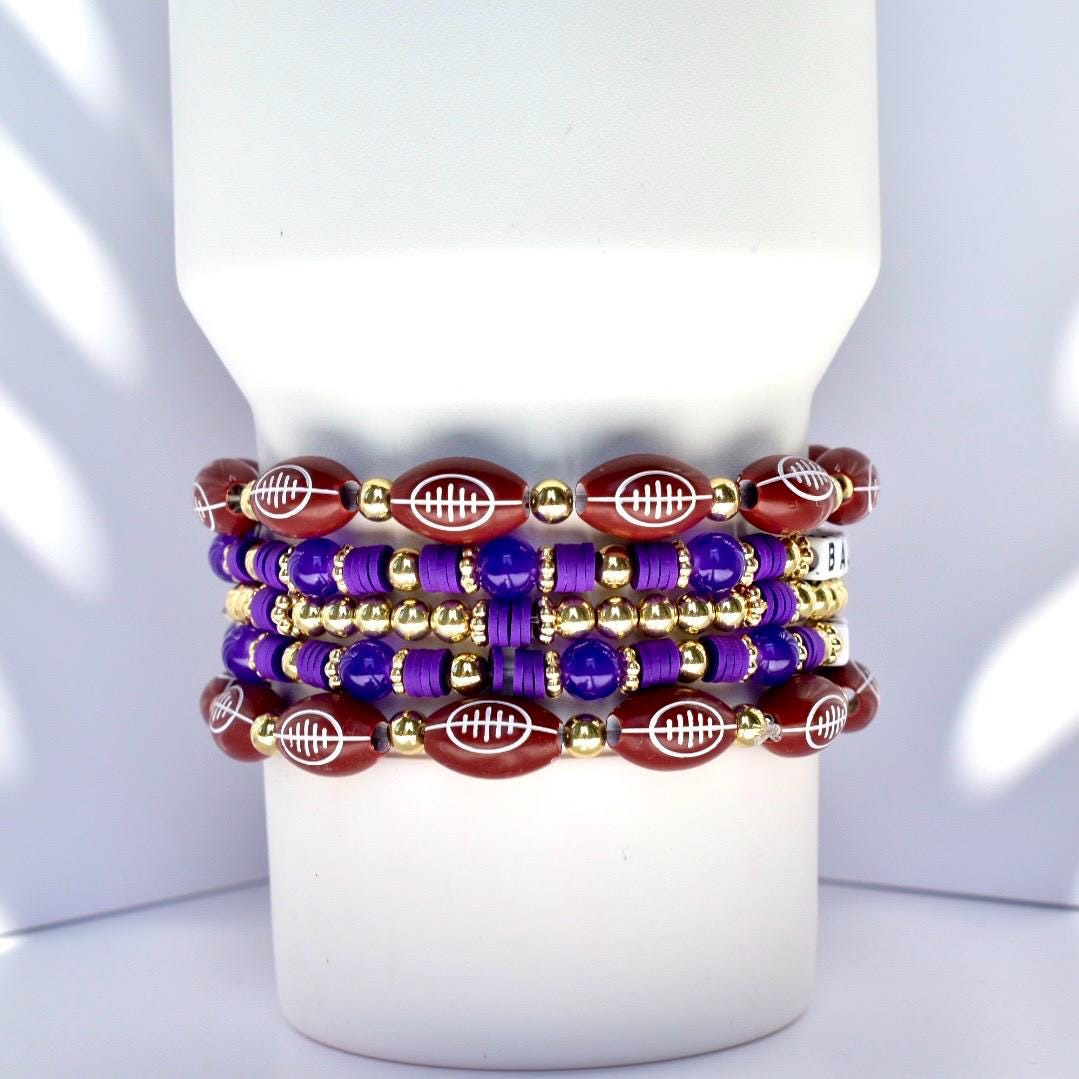 Stack of beaded bracelets in shades of burgundy, purple, and gold. The bracelets feature football-shaped beads, round gold beads, and purple beads. The bracelets are stacked on a white surface.
