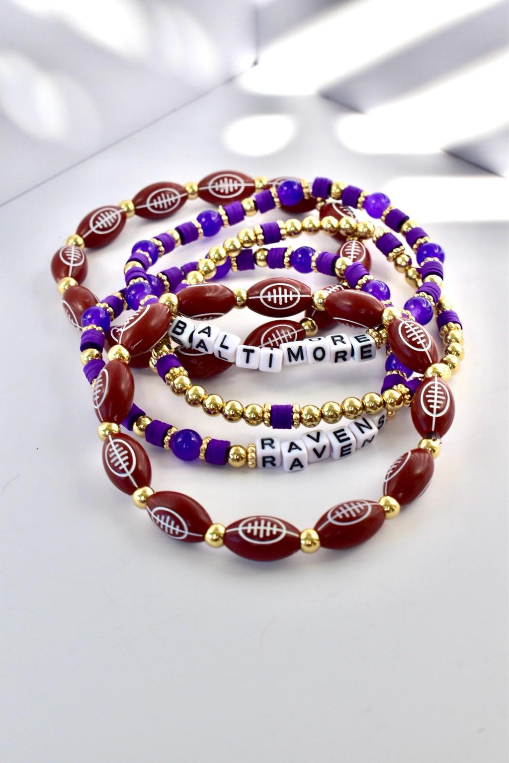 Several bracelets with a football theme. The bracelets are made with maroon football-shaped beads, gold beads, and purple beads. Some bracelets spell out "BALTIMORE RAVENS" with white letter beads.