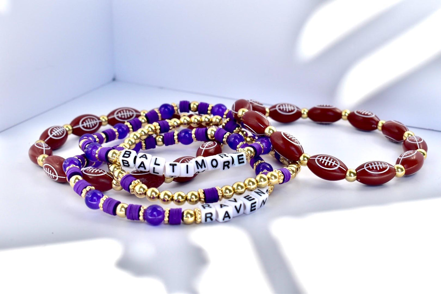 Several beaded bracelets in purple, gold, and burgundy. Some beads are football-shaped, others are round or square. One bracelet spells out "BALTIMORE RAVEN" in white letter beads.
