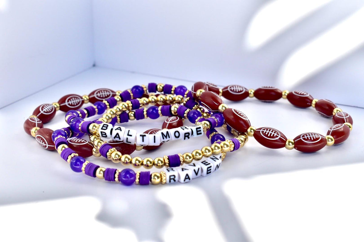 Several beaded bracelets in purple, gold, and burgundy. Some beads are football-shaped, others are round or square. One bracelet spells out "BALTIMORE RAVEN" in white letter beads.