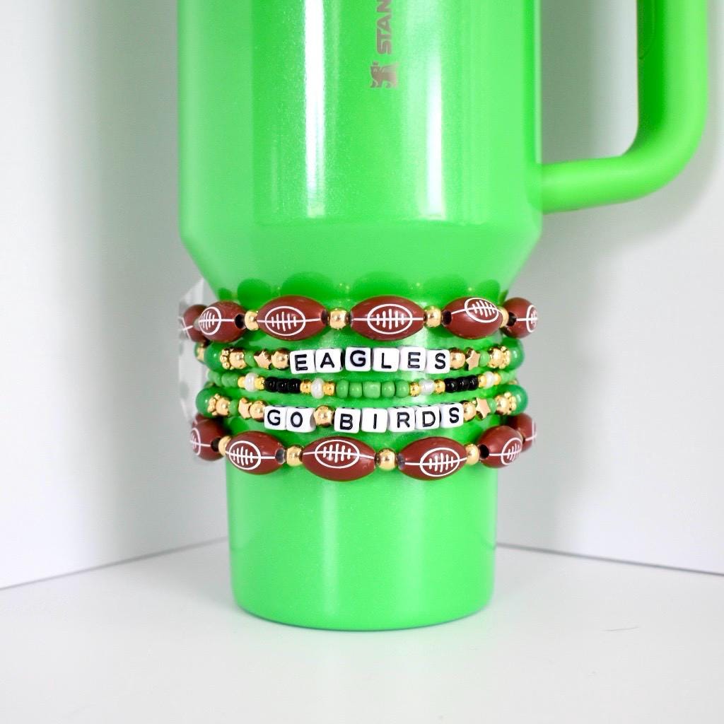 A green Stanley mug with several beaded bracelets. The bracelets feature brown football-shaped beads, gold beads, and white letter beads spelling "EAGLES" and "GO BIRDS".
