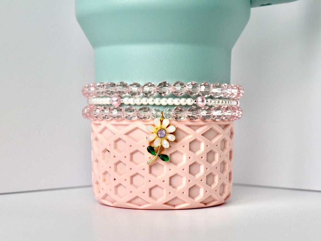 A light pink, geometric patterned container holds three bracelets and a flower charm. The bracelets are made of clear, faceted beads and white pearls. The flower charm has a yellow center, purple accent, and green stem. The container is light pink and has a mint green top.