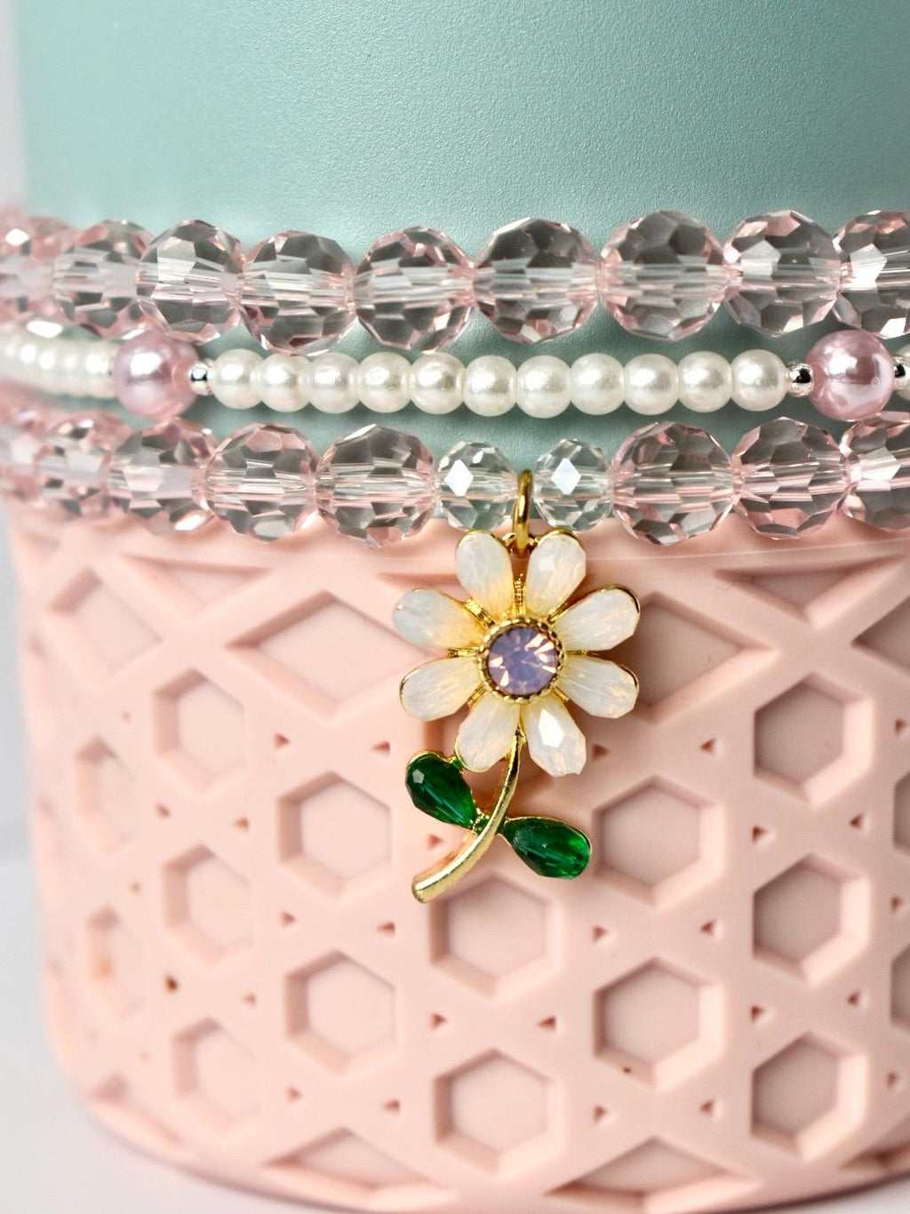 A close-up of a beaded bracelet set with a flower charm. The bracelets feature clear, pink, and white beads. The flower charm is gold with white petals, a purple center, and green leaves. The bracelets are displayed on a pink, textured surface.