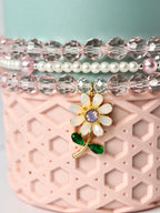 A close-up of a beaded bracelet set with a flower charm. The bracelets feature clear, pink, and white beads. The flower charm is gold with white petals, a purple center, and green leaves. The bracelets are displayed on a pink, textured surface.