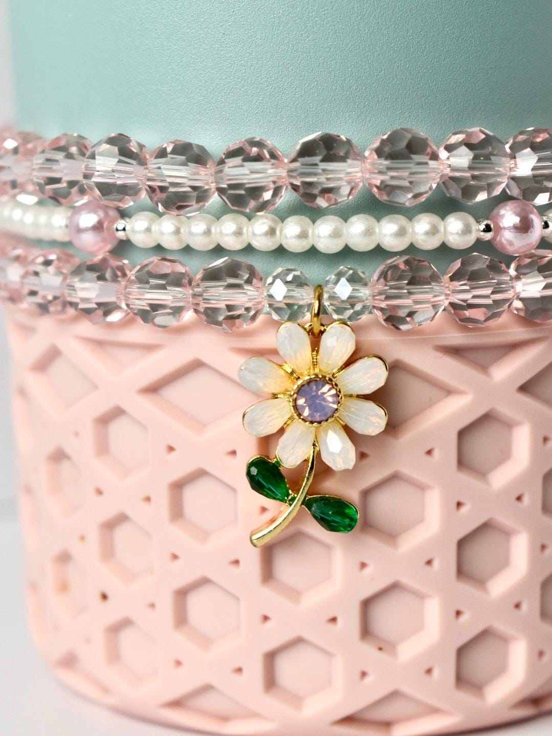 A close-up of a beaded bracelet set with a flower charm. The bracelets feature clear, pink, and white beads. The flower charm is gold with white petals, a purple center, and green leaves. The bracelets are displayed on a pink, textured surface.