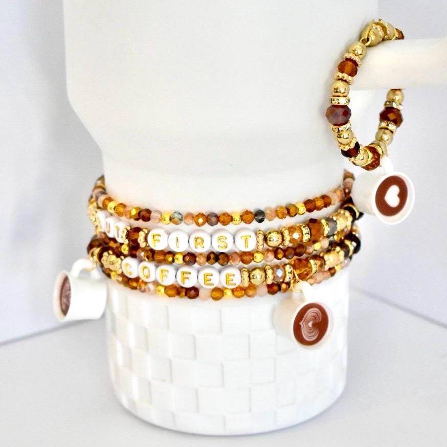 A stack of five Stanley bracelets in amber, brown, and gold with coffee cup charms and letter beads that say ‘BUT FIRST COFFEE.’ Also pictured is a matching handle bracelet with the same color beads and a coffee cup charm hanging from the mini bracelet.