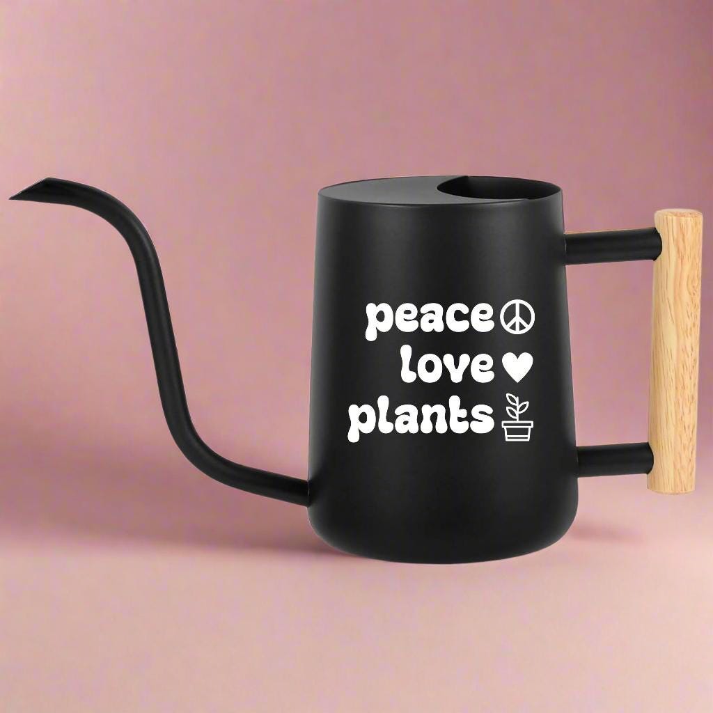Black metal watering can with a light brown wood handle with the words peace love plants in white vinyl on the side of the can along with a peace sign heart and plant icon.