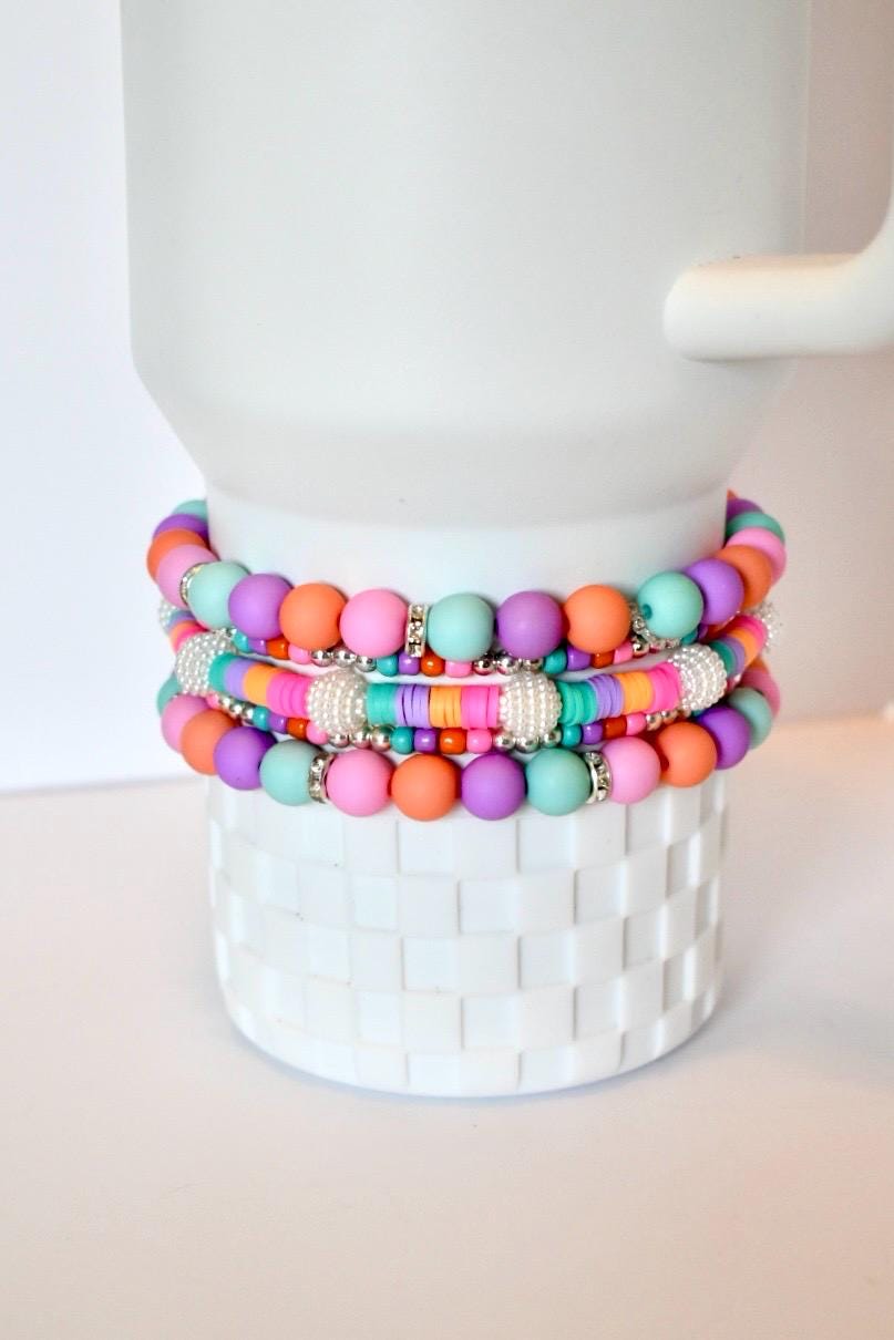 A stack of colorful beaded bracelets in shades of pink, orange, purple, and teal. The bracelets are accented with silver-colored beads and are displayed on a white, textured surface.