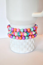 A stack of colorful beaded bracelets in shades of pink, orange, purple, and teal. The bracelets are accented with silver-colored beads and are displayed on a white, textured surface.