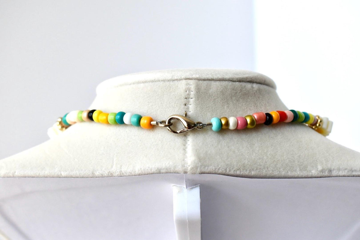 A necklace featuring a strand of small, multi-colored beads in shades of yellow, teal, white, pink, black, and gold. The necklace is secured with a silver-toned metal lobster claw clasp, which is positioned at the center of the strand.