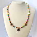 A beaded necklace featuring a vibrant array of multicolored small beads, two gold-toned bee charms, two white flower-shaped beads, and a central circular gold-toned pendant engraved with the capital letter M in a serif font.