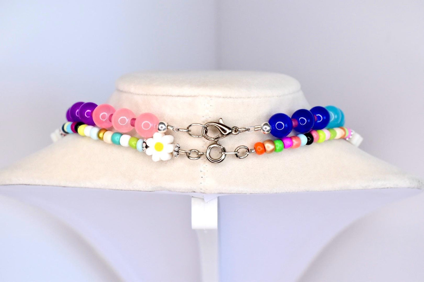Two colorful beaded necklaces displayed on a white surface. The top necklace features purple, pink, and white beads with a daisy charm. The bottom necklace has blue, green, orange, and pink beads. Both necklaces have silver clasps.