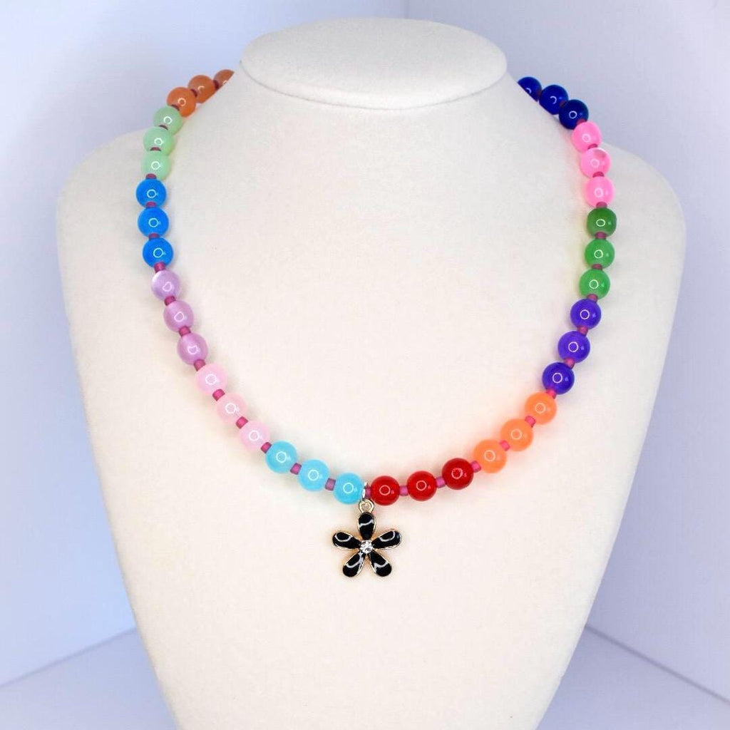 A colorful beaded necklace displayed on a white mannequin. The necklace features a gradient of round beads in various colors, including red, orange, yellow, green, blue, purple, and pink. A black flower pendant hangs at the center.