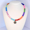 A colorful beaded necklace displayed on a white mannequin. The necklace features a gradient of round beads in various colors, including red, orange, yellow, green, blue, purple, and pink. A black flower pendant hangs at the center.