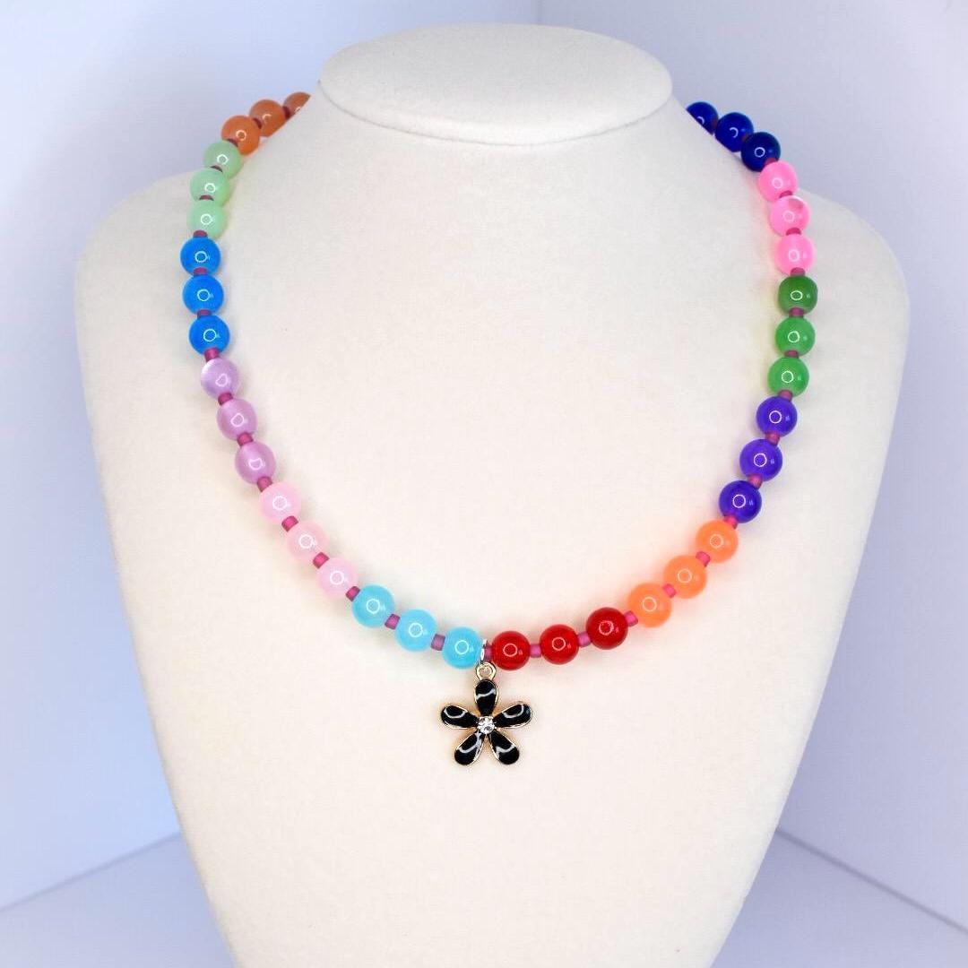 A colorful beaded necklace displayed on a white mannequin. The necklace features a gradient of round beads in various colors, including red, orange, yellow, green, blue, purple, and pink. A black flower pendant hangs at the center.
