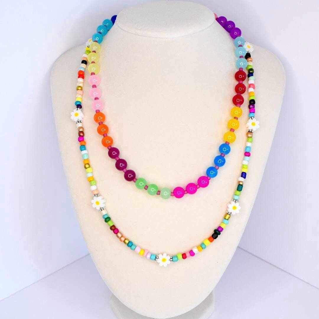 A colorful beaded necklace with a rainbow color scheme. The necklace features large, round beads in various colors, including blue, green, pink, orange, and purple. Smaller beads and white daisy accents are interspersed throughout the design, creating a playful and vibrant accessory.