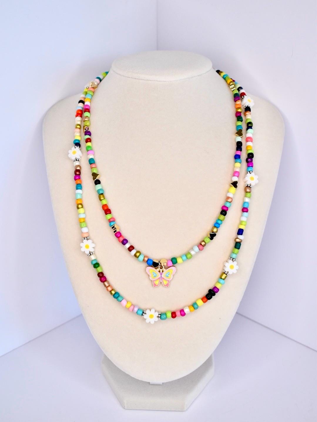 Two colorful beaded necklaces displayed on a mannequin. The necklaces feature a variety of small, round beads in multiple colors, including red, yellow, green, blue, and black. One necklace has a butterfly charm, and both have white daisy accents. The necklaces are layered, creating a vibrant and playful look.