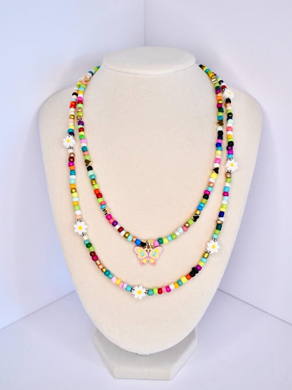 Two colorful beaded necklaces displayed on a mannequin. The necklaces feature a variety of small, round beads in multiple colors, including red, yellow, green, blue, and black. One necklace has a butterfly charm, and both have white daisy accents. The necklaces are layered, creating a vibrant and playful look.